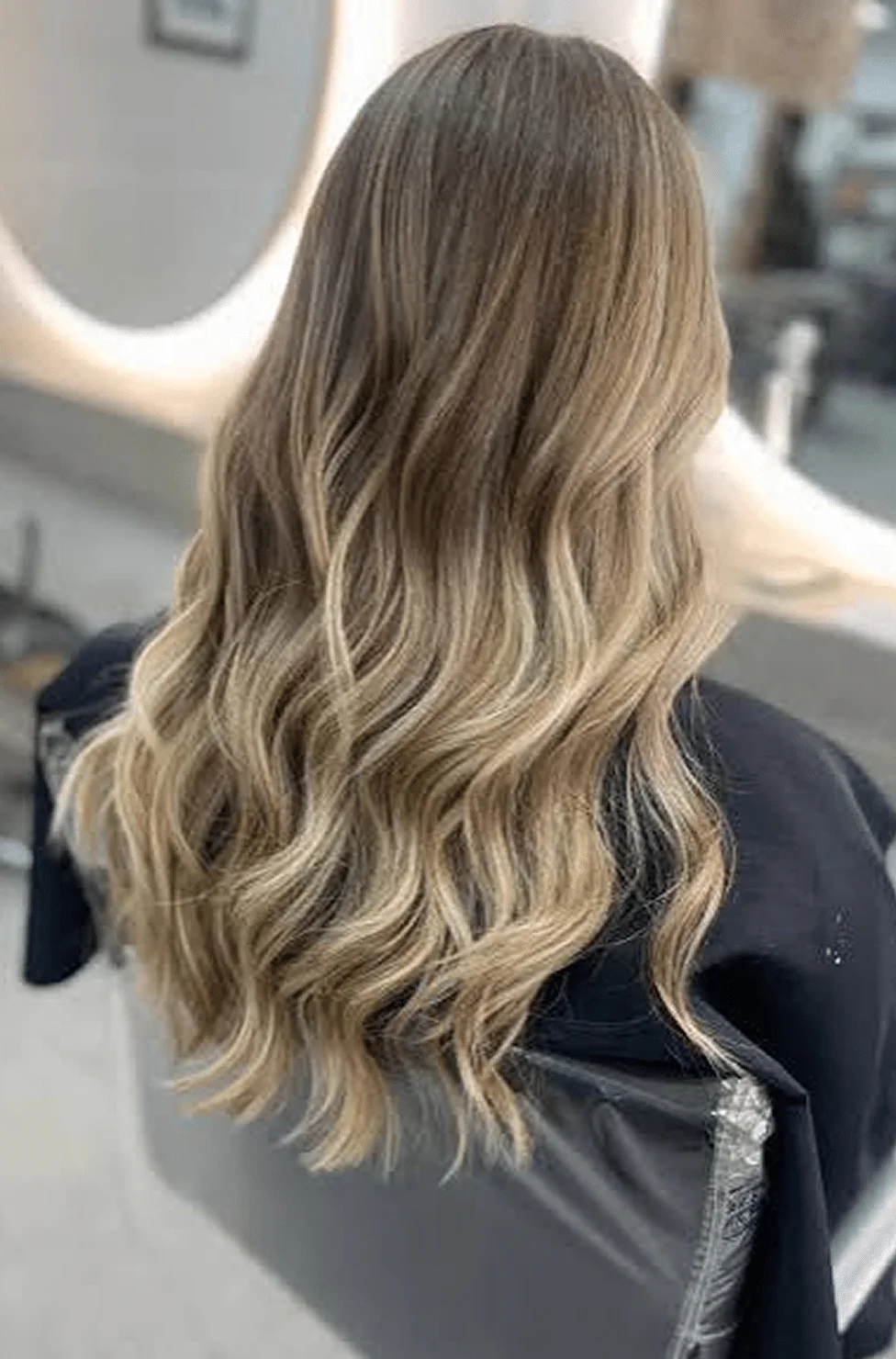 Back view of a person with shoulder-length blonde hair in loose waves at a hair salon.