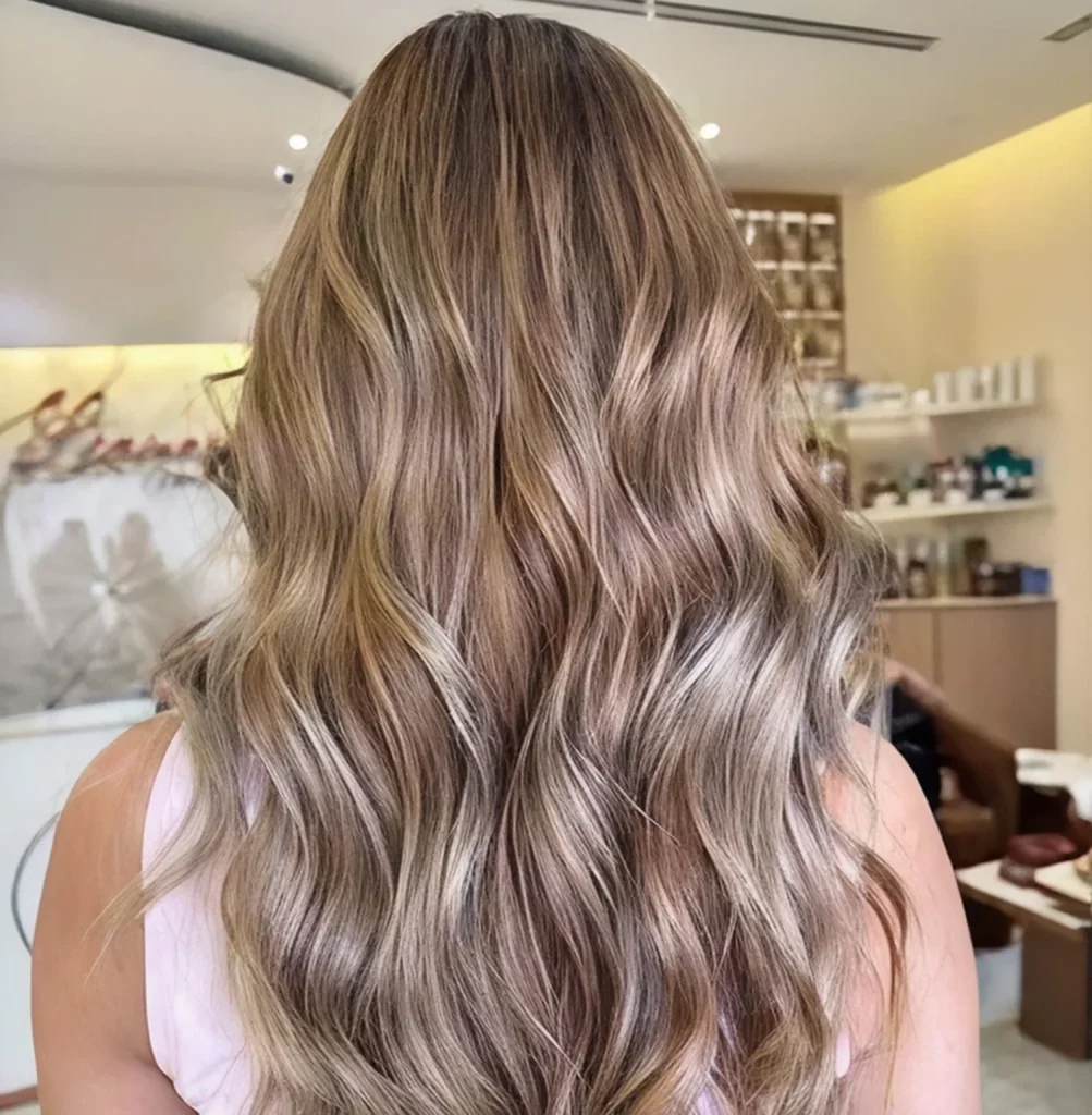Back view of a woman with long, wavy blonde balayage hair in a salon.