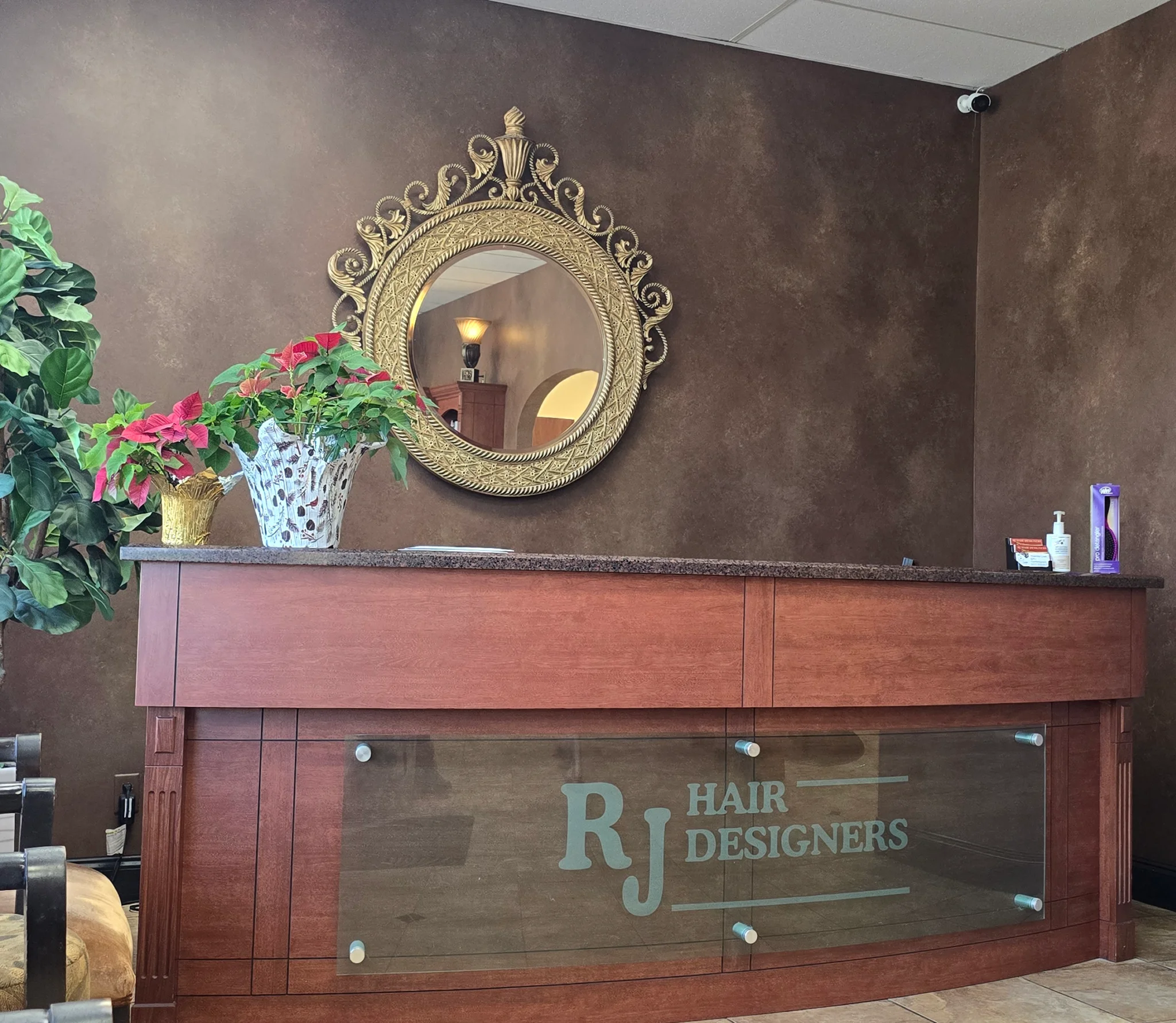 Decorative gold oval mirror on a dark brown textured wall above a wooden reception counter with green plants nearby.
