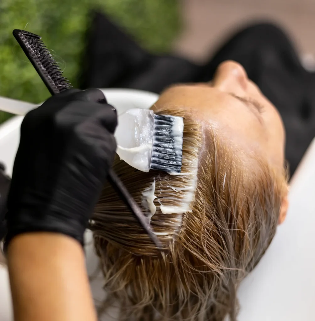 Person applying hair dye to a client's hair with a brush, wearing black gloves and using a comb nearby