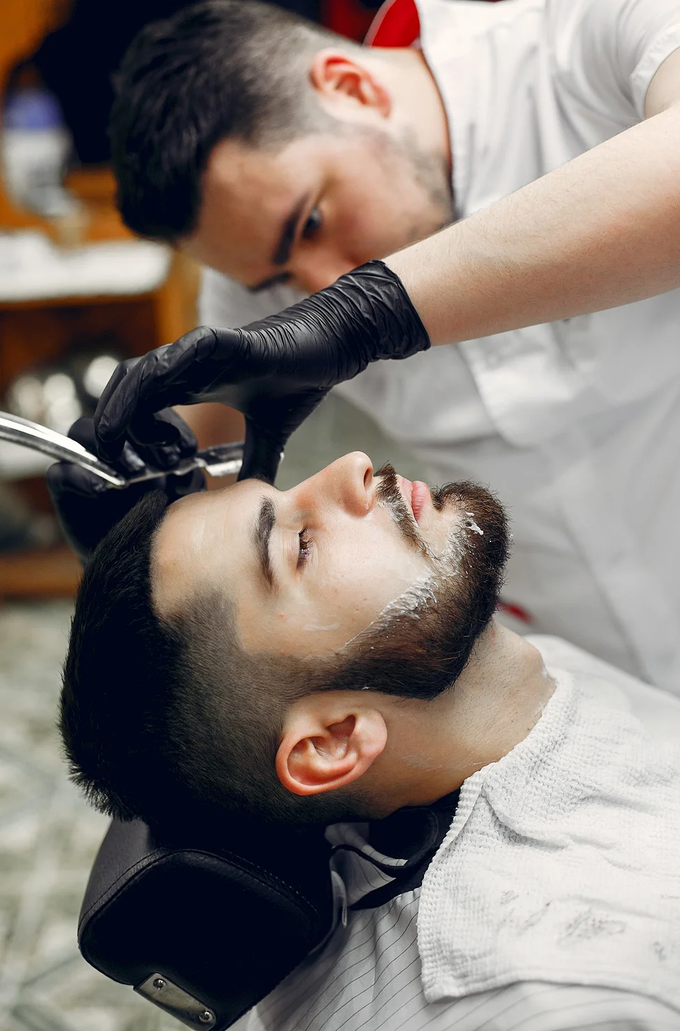 Bearded man reclining in a barber chair as a barber with black gloves shaves his face with a straight razor and shaving foam nearby.