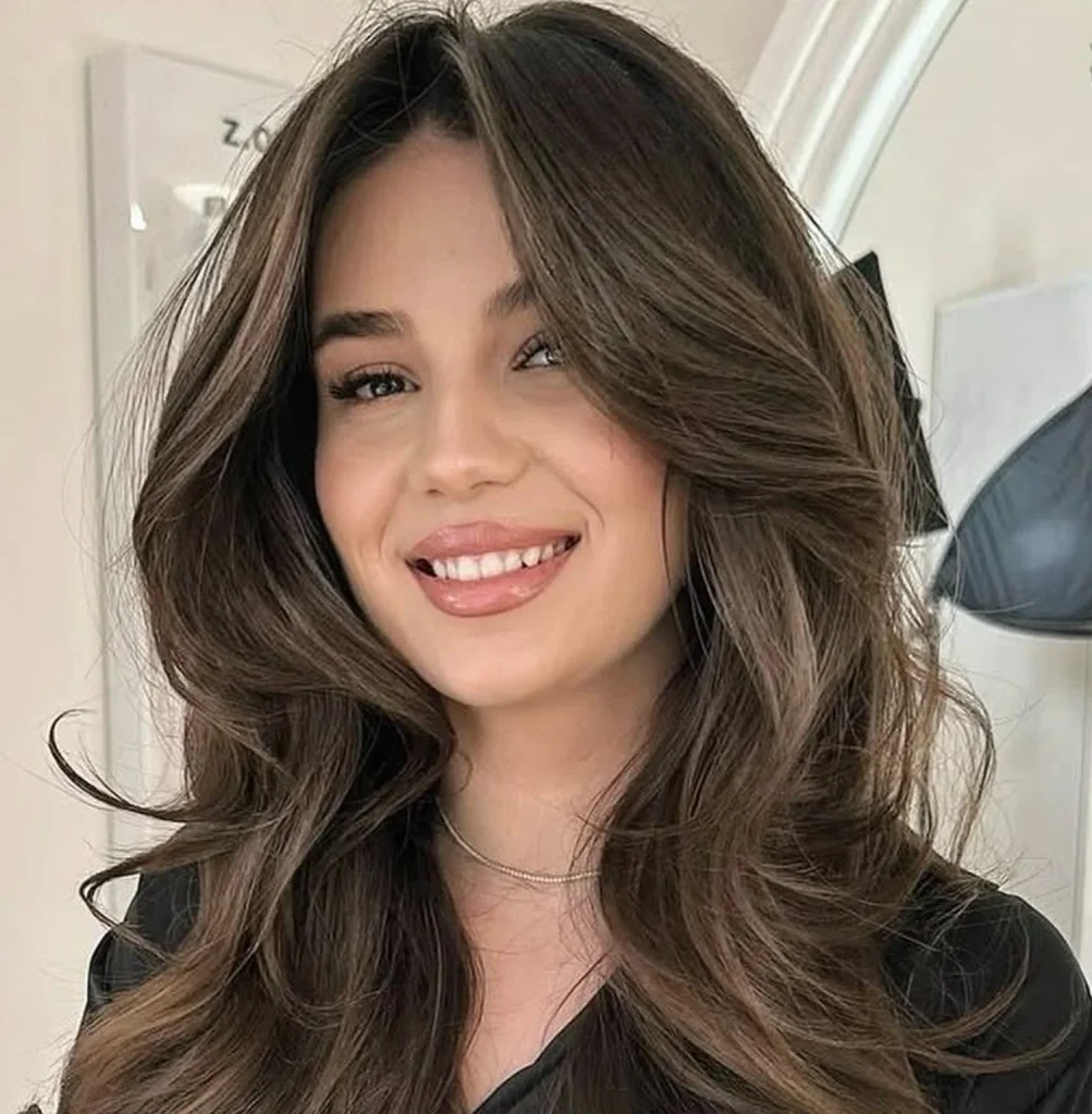 Close-up portrait of a smiling woman with long, wavy brown hair wearing a black top in a bright indoor setting.