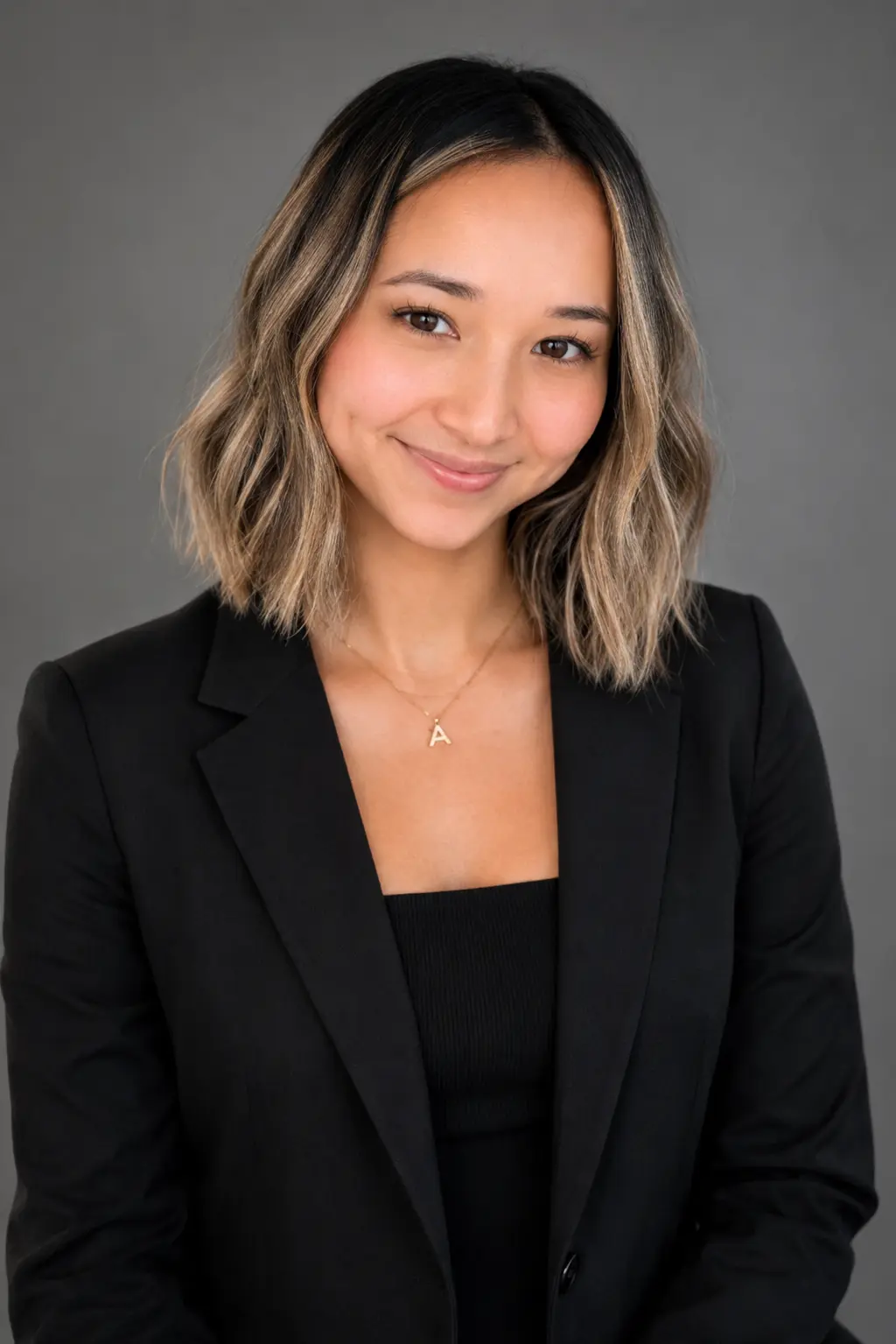 Smiling woman in a black blazer and top, shoulder-length wavy hair, wearing a gold A pendant against a gray backdrop.