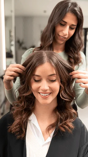 Hair stylist styling a woman's brown wavy hair in a salon; client wears a white blouse and black cape.
