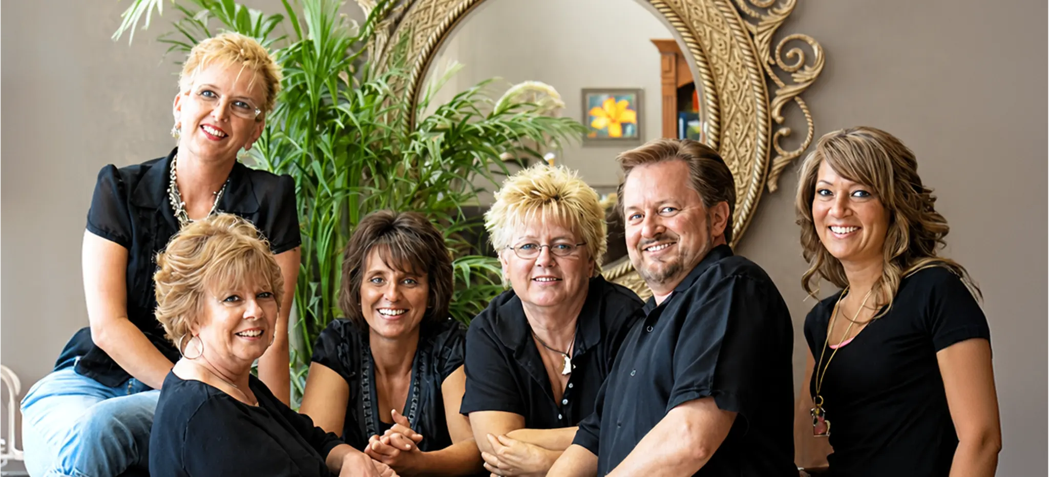 Group of six adults (five women, one man) wearing black, posing and smiling in front of a decorative mirror and plants.