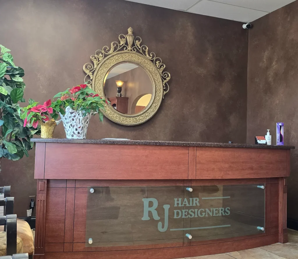 Reception desk with glass panel labeled RJ Hair Designers, brown textured wall, a large ornate round mirror, and potted plants nearby