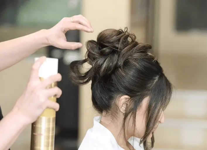 Stylist fixes a woman's dark hair into a voluminous updo while spraying with hairspray in a salon.