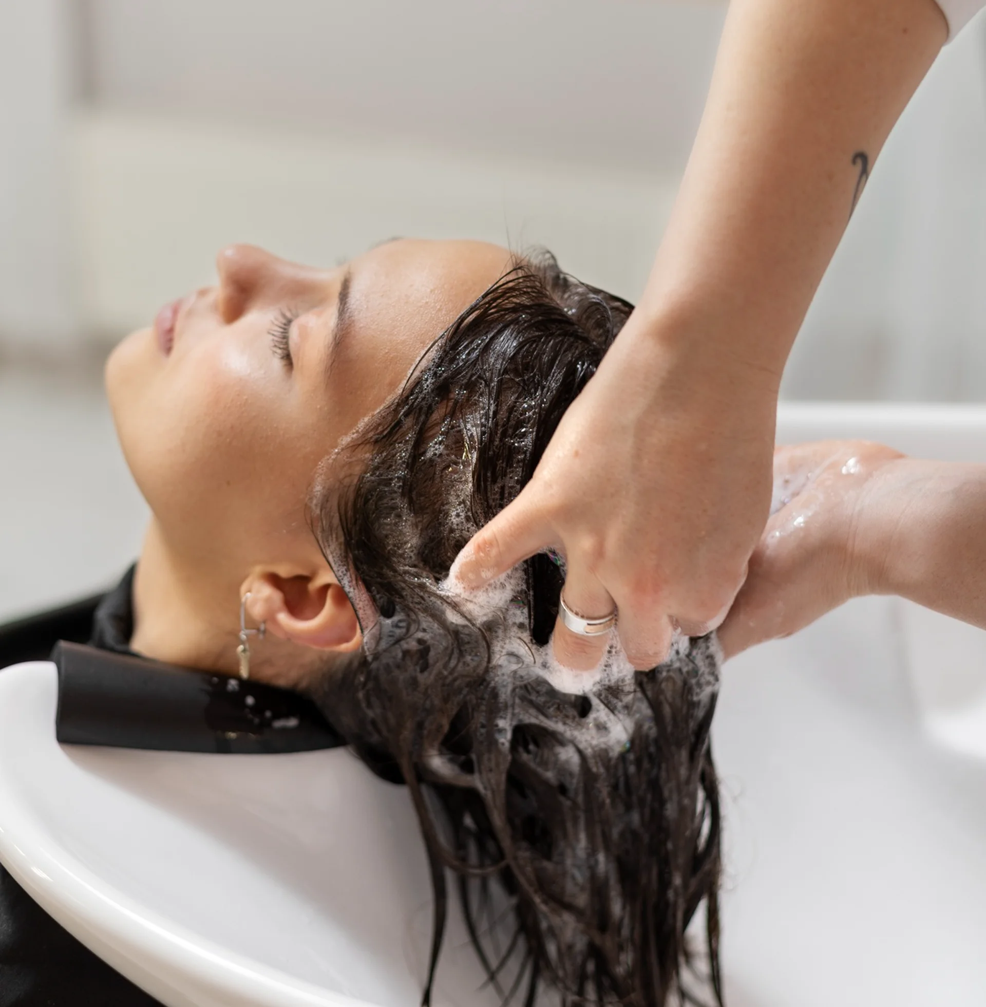 Person lying back over a sink while shampooing wet hair with foam around and arm raised above head.